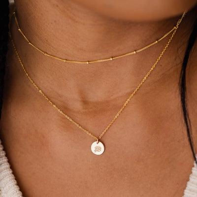 A close-up of a person wearing a thin gold choker with stationary round beads layered with a delicate gold pendant chain featuring a flat round gold disc. The disc is engraved with a peony floral motif.