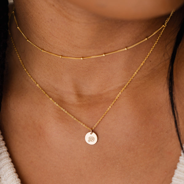 A close-up of a person wearing a thin gold choker with stationary round beads layered with a delicate gold pendant chain featuring a flat round gold disc. The disc is engraved with a peony floral motif.