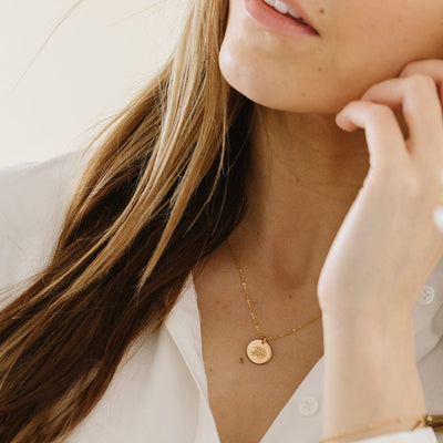 A close-up of a woman in a white button up top wearing a delicate gold pendant chain with a round flat disc charm, engraved with a peony flower.