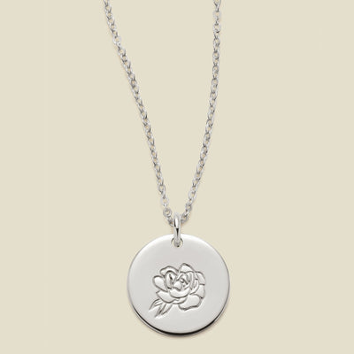 A silver chain necklace with a round silver disc pendant featuring an engraved peony flower design, displayed against a solid light beige background.