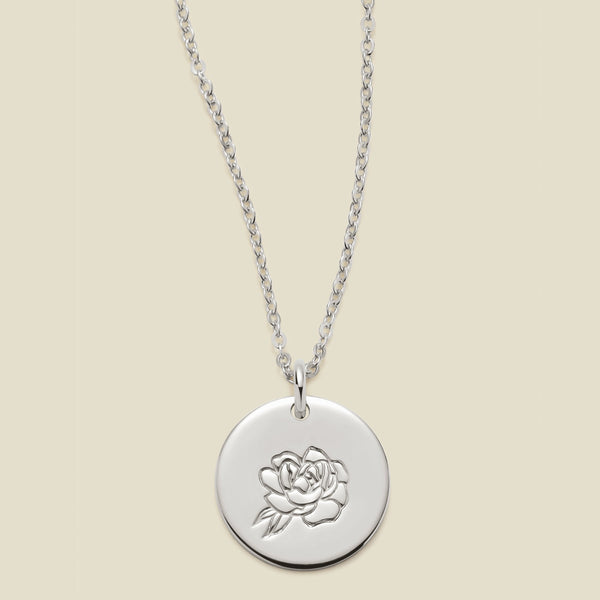 A silver chain necklace with a round silver disc pendant featuring an engraved peony flower design, displayed against a solid light beige background.
