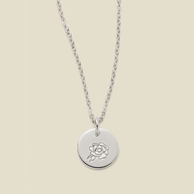 A silver chain necklace with a round silver disc pendant featuring an engraved peony flower design, displayed against a solid light beige background.