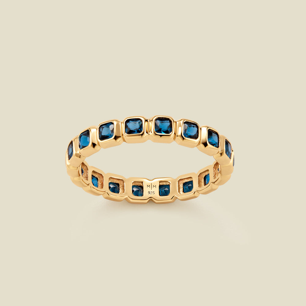 A gold eternity ring featuring a continuous row of square cut dark blue gemstones, shown on a solid light beige background.