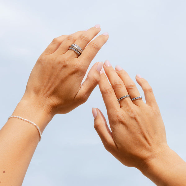 A close-up of two hands wearing multiple stacked silver rings, with some of the rings having different colors of square cut gemstones, and a matching silver bracelet.