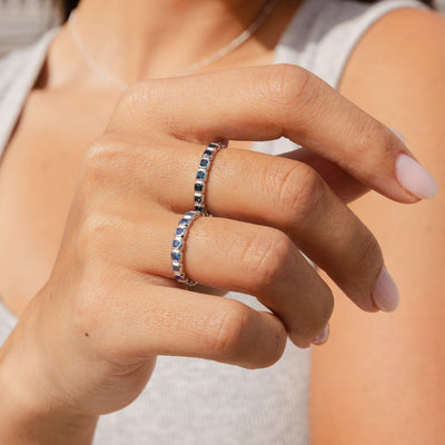 A close-up of a person's hand wearing two silver eternity rings, each with a different shade of blue square cut gemstones in a single continuous row.