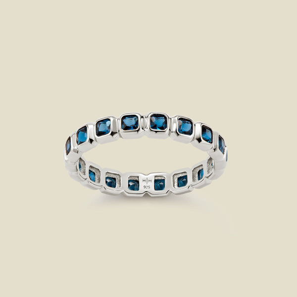 A silver eternity ring featuring a continuous row of square cut dark blue gemstones, shown on a solid light beige background.