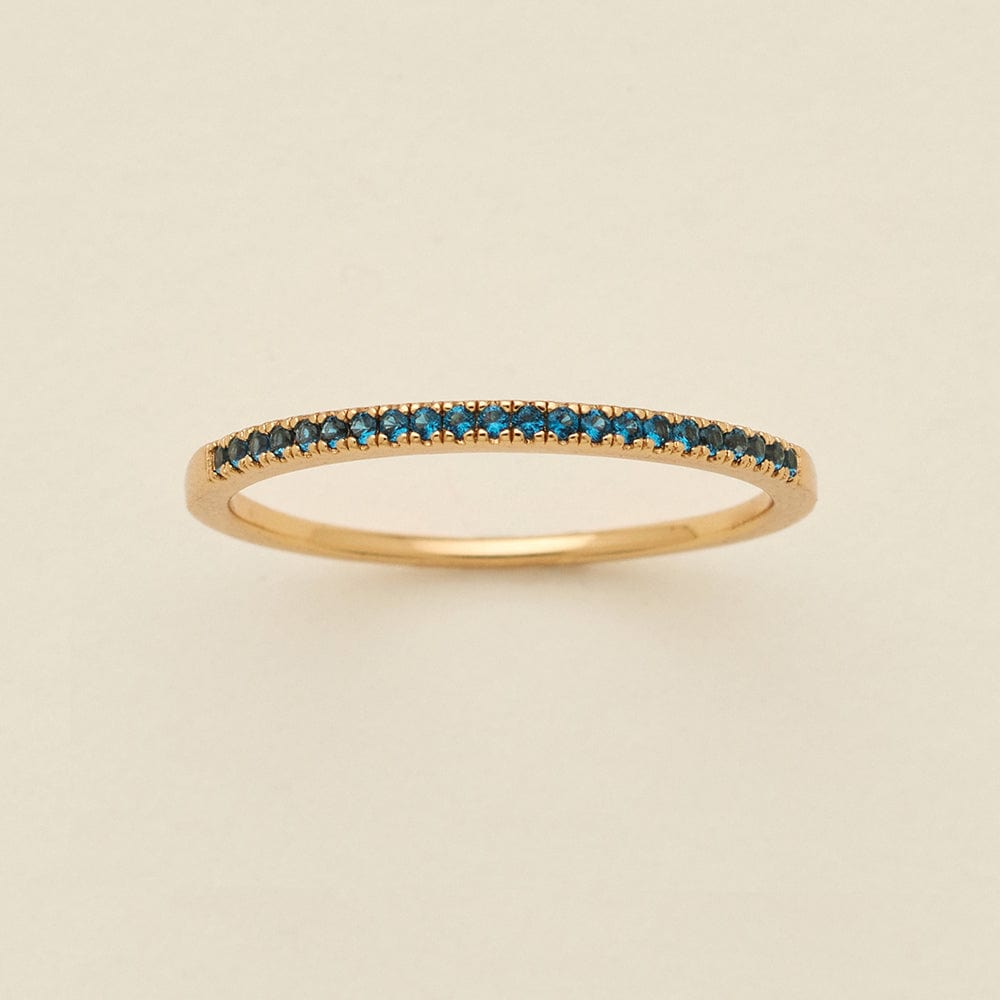 A gold ring featuring a single row of small round-cut dark blue-green gemstones, photographed on a clean, solid, warm off-white background.