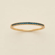 A gold ring featuring a single row of small round-cut dark blue-green gemstones, photographed on a clean, solid, warm off-white background.
