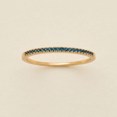 A gold ring featuring a single row of small round-cut dark blue-green gemstones, photographed on a clean, solid, warm off-white background.
