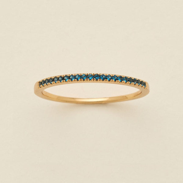 A gold ring featuring a single row of small round-cut dark blue-green gemstones, photographed on a clean, solid, warm off-white background.