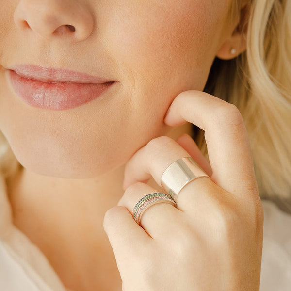 A close-up of a hand resting against a person's cheek, showcasing a thick polished gold cigar band ring and four stacked thin band rings, each with a single row of gemstones in the colors blue-green, emerald green, red, and white.