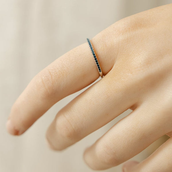 A close-up of a hand with a thin silver ring, featuring a single row of small dark blue-green gemstones, set against a light beige backdrop.