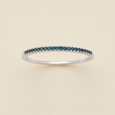 A silver ring featuring a single row of small round-cut dark blue-green gemstones, photographed on a clean, solid, warm off-white background.