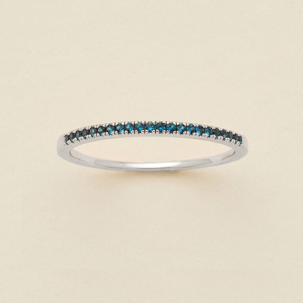 A silver ring featuring a single row of small round-cut dark blue-green gemstones, photographed on a clean, solid, warm off-white background.