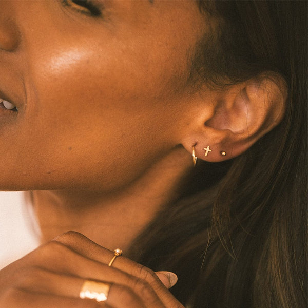 A close-up of a woman's ear wearing a small gold hoop, a gold cross stud and a gold round bead stud.