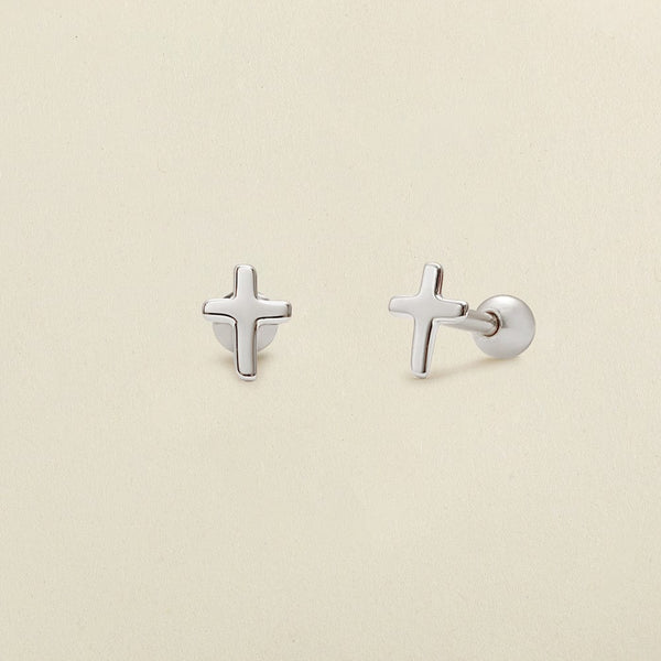 A pair of minimalistic silver cross stud earrings with round ball backings, shown on a solid light beige background.