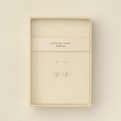 Two pairs of silver stud earrings displayed inside a cream-colored jewelry box. One earring is a small round dot, and the other is a small cross.
