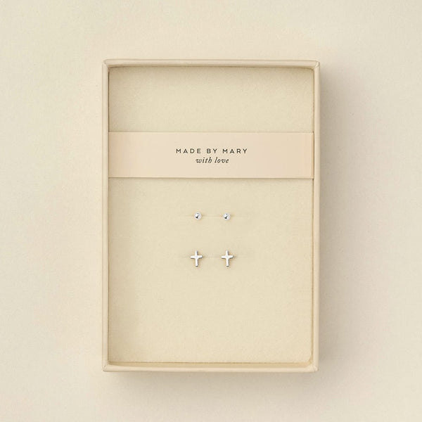 Two pairs of silver stud earrings displayed inside a cream-colored jewelry box. One earring is a small round dot, and the other is a small cross.