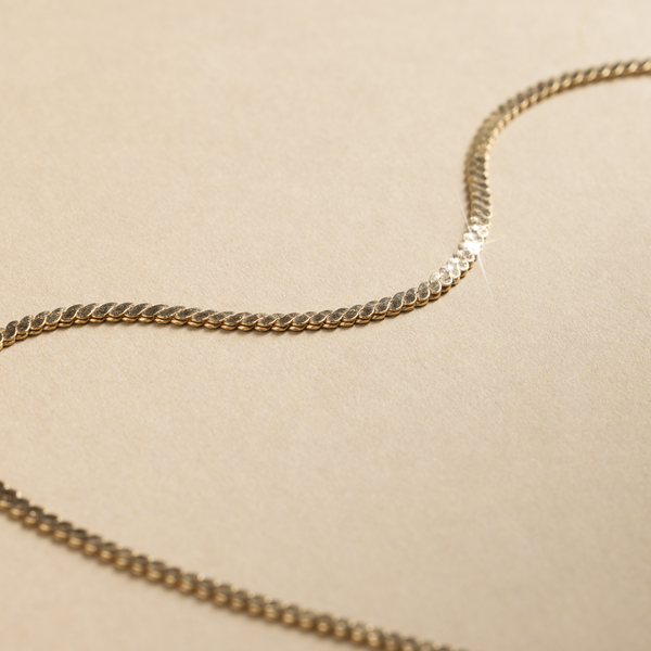 A gold, flat braided chain necklace set against a neutral textured background.