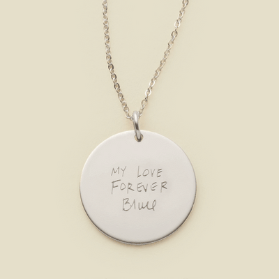 A dainty silver chain necklace with a round silver pendant engraved with handwritten script.