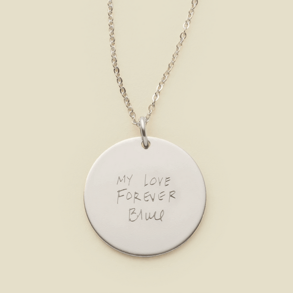 A dainty silver chain necklace with a round silver pendant engraved with handwritten script.