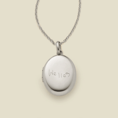 A dainty silver chain necklace with a silver oval locket pendant engraved with handwritten script.
