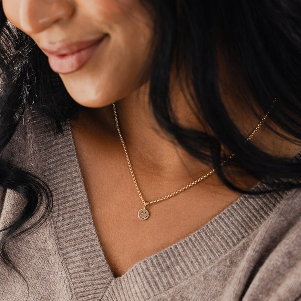 A close-up of a person in a grey v-neck knit top, paired with a gold chain necklace featuring a flat polished round smiley face charm.