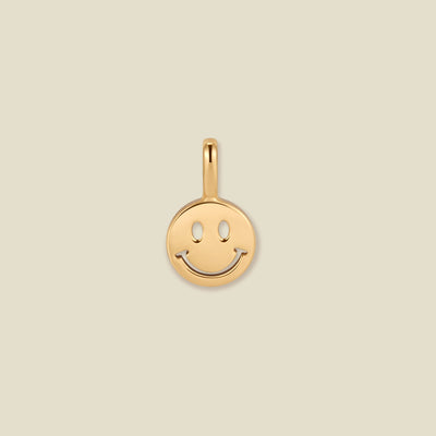 A small round gold smiley face charm with a polished finish and a top loop, centered on a solid light beige background.