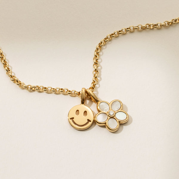 A close-up of a gold circular link chain with a gold flat round smiley face charm and a gold daisy charm with a white inlay on the petals and a clear accent stone in the center, shown on a light beige background.
