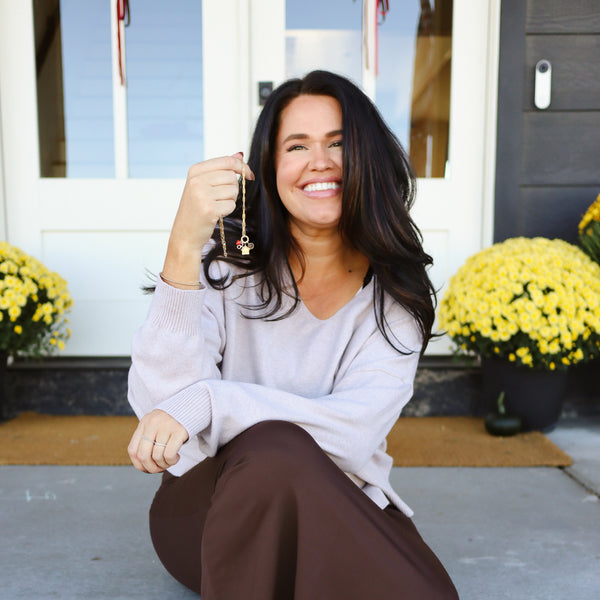 A smiling woman sitting on a porch step with pots of yellow flowers in the background, wearing a loose grey sweater top and brown pants. She is holding and dangling a gold chain necklace with multiple charms on a round ring in the center.