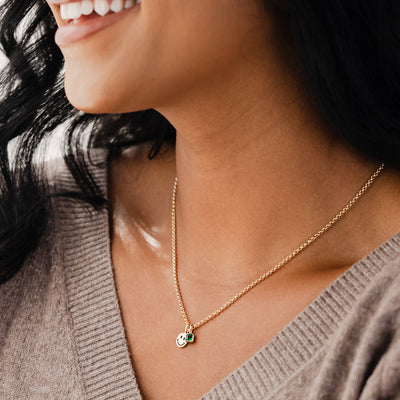 A close-up of a person in a grey v-neck knit sweater, wearing a gold chain necklace with a gold flat round smiley face charm and a square green gemstone charm.