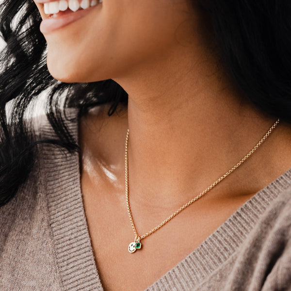 A close-up of a person in a grey v-neck knit sweater, wearing a gold chain necklace with a gold flat round smiley face charm and a square green gemstone charm.
