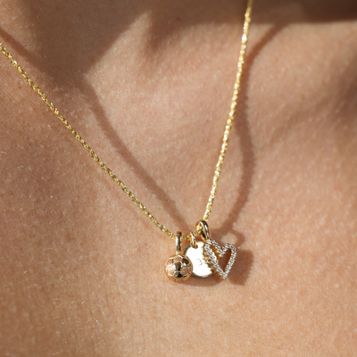 A close-up of a person wearing a textured gold chain necklace with a gold soccer ball charm, a gold flat round disc charm, and a gold heart outline charm with clear gemstones.