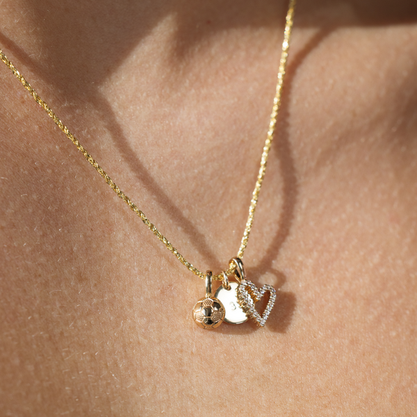 A close-up of a person wearing a textured gold chain necklace with a gold soccer ball charm, a gold flat round disc charm, and a gold heart outline charm with clear gemstones.