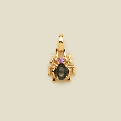 A gold spider pendant featuring a large black oval stone and a small pink gemstone on its back, centered on a light beige background.