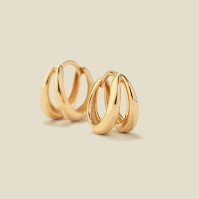 Two polished gold huggie hoop earrings made up of two stationary separate hoops attached at the earring post, shown on a warm beige background, with one earring facing forward and the other angled to show the side profile.