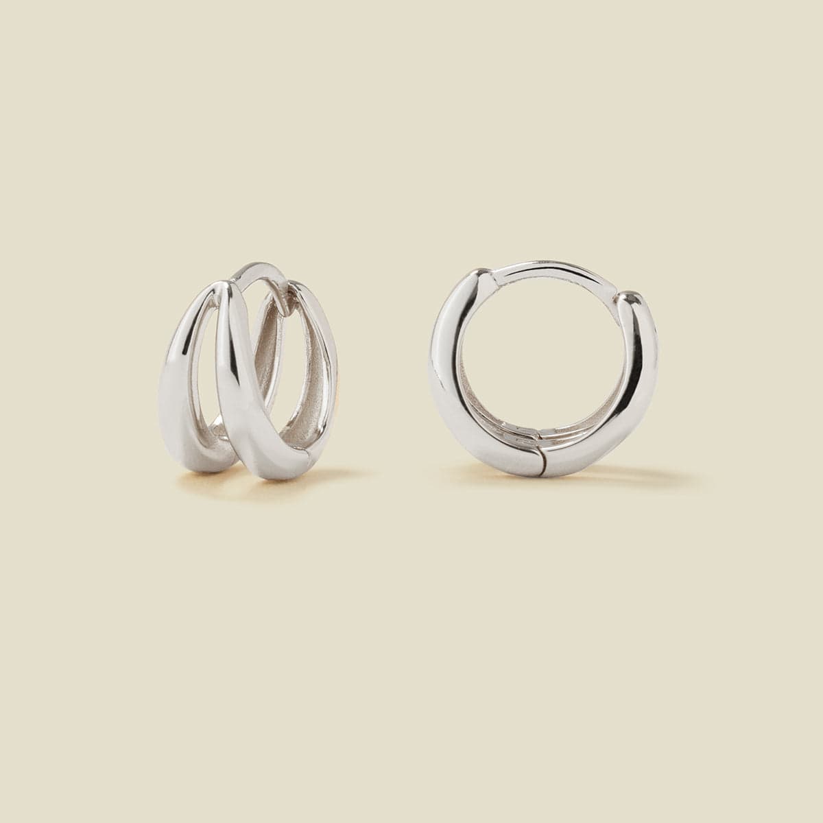 A pair of polished silver huggie hoop earrings with a split down the center shown on a warm beige background. One earring is shown from a side angle, revealing its layered design, and the other is shown from a front angle.