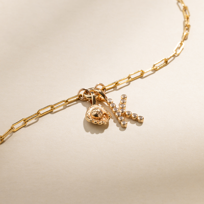 A gold paperclip link charm bracelet featuring a gold baseball mitt charm, and a gold initial 'K' charm with clear gemstones, set against a beige background with natural shadows.