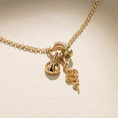 A gold chain necklace with a round, link lock charm holder with clear gemstones, featuring a gold basketball charm, a gold snake charm, and a gold ring charm comprised of square, bezel-set green stones, set against a beige background with shadows.