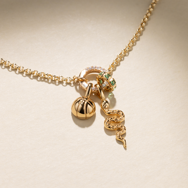 A gold chain necklace with a round, link lock charm holder with clear gemstones, featuring a gold basketball charm, a gold snake charm, and a gold ring charm comprised of square, bezel-set green stones, set against a beige background with shadows.