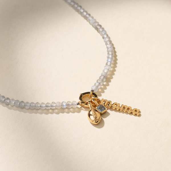 A beaded grey gemstone necklace with a gold, round charm holder in the center, featuring a gold football charm, a square, light blue bezel-set gemstone charm, and a script font vertical charm with the word 'mama', set against a solid beige background.