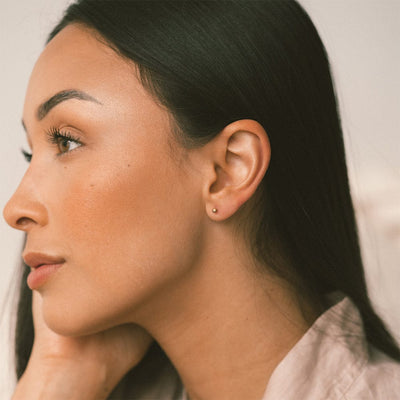 A side profile of a woman with dark hair wearing a small round gold bead stud, shown against a solid light beige background.