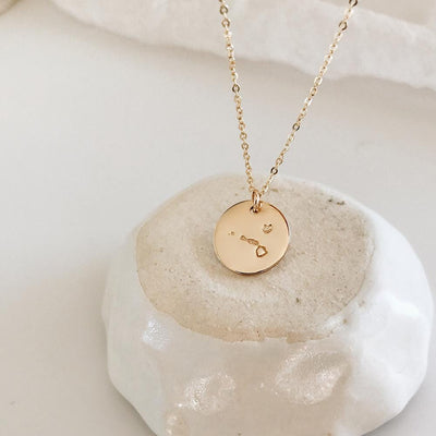 A delicate gold pendant chain necklace with a round flat disc engraved with the outline of Hawaii, resting on a white and beige rock with a white background.