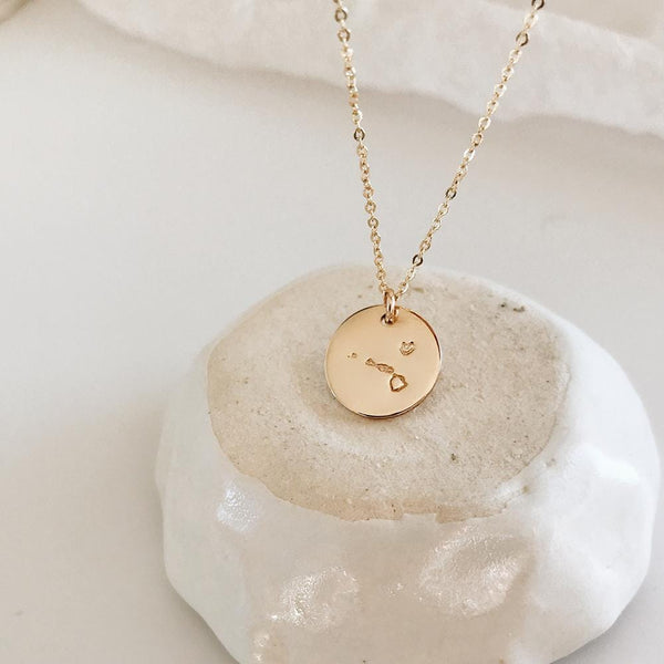 A delicate gold pendant chain necklace with a round flat disc engraved with the outline of Hawaii, resting on a white and beige rock with a white background.