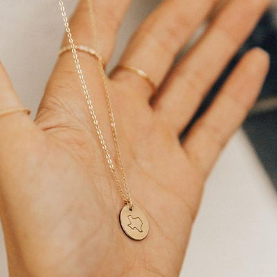 A person's hand holding a delicate gold pendant chain necklace with a round disc engraved with the outline of the state of Texas.