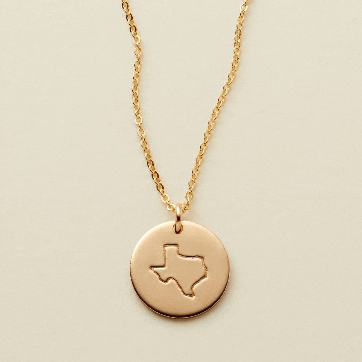 A gif of a delicate gold pendant chain necklace with a round disc charm, changing engravings between outlines of different states.
