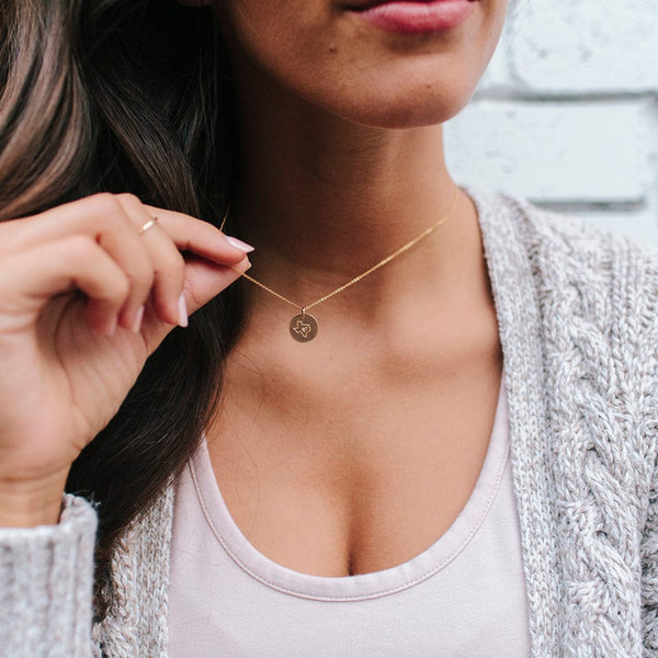 A woman in a white top and a grey knit cardigan holding a gold pendant necklace off her neck, showcasing the round flat disc charm engraved with the outline of the state of Texas with a heart in the center.