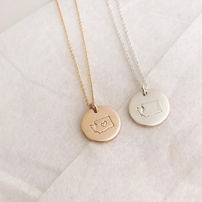 A gold and a silver pendant chain necklace, each with a round disc charm with the outline of the state of Washington. The gold charm has a heart outline in the center of the state.