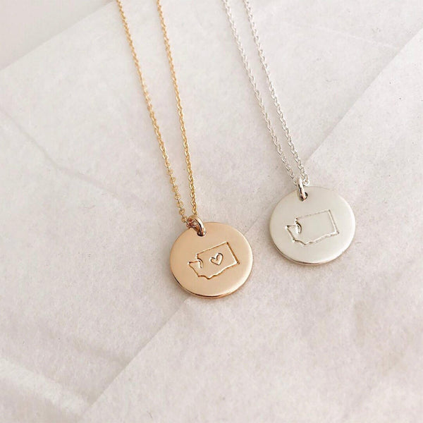 A gold and a silver pendant chain necklace, each with a round disc charm with the outline of the state of Washington. The gold charm has a heart outline in the center of the state.