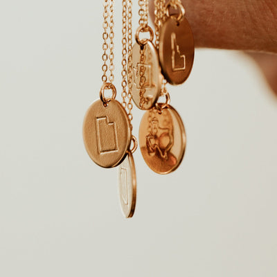 A close-up of five round discs hanging from gold chain necklaces, each engraved with a different outline of a state.